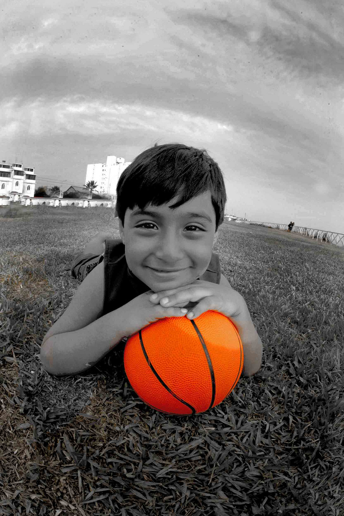 FOografia en blanco y nego a excepcion del balón de baloncesto de color naraja que tiene el niño entre sus manos. El nio eta tumbado boca a bajo en el cesped mirando sonriente a cámara.